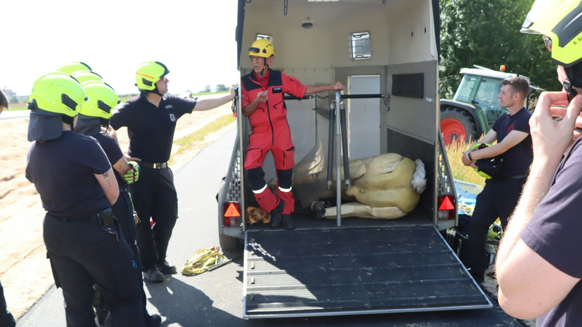 Auf dem Bild ist ein Pferdanhänger zu sehen, in dem der Pferde-Dummy auf der Seite liegt. Feuerwehrleute und der Seminarleiter stehen drumherum.