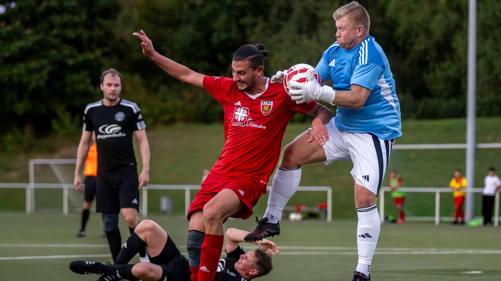 Das Bild zeigt den Torwart und einen Gegenspieler im Zweikampf. Hinter ihnen liegt ein weiterer Akteur am Boden.