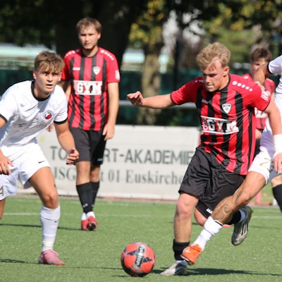 Das Bild zeigt einen Zweikampf zweier Spieler um den Ball. Weitere Akteure schauen zu.