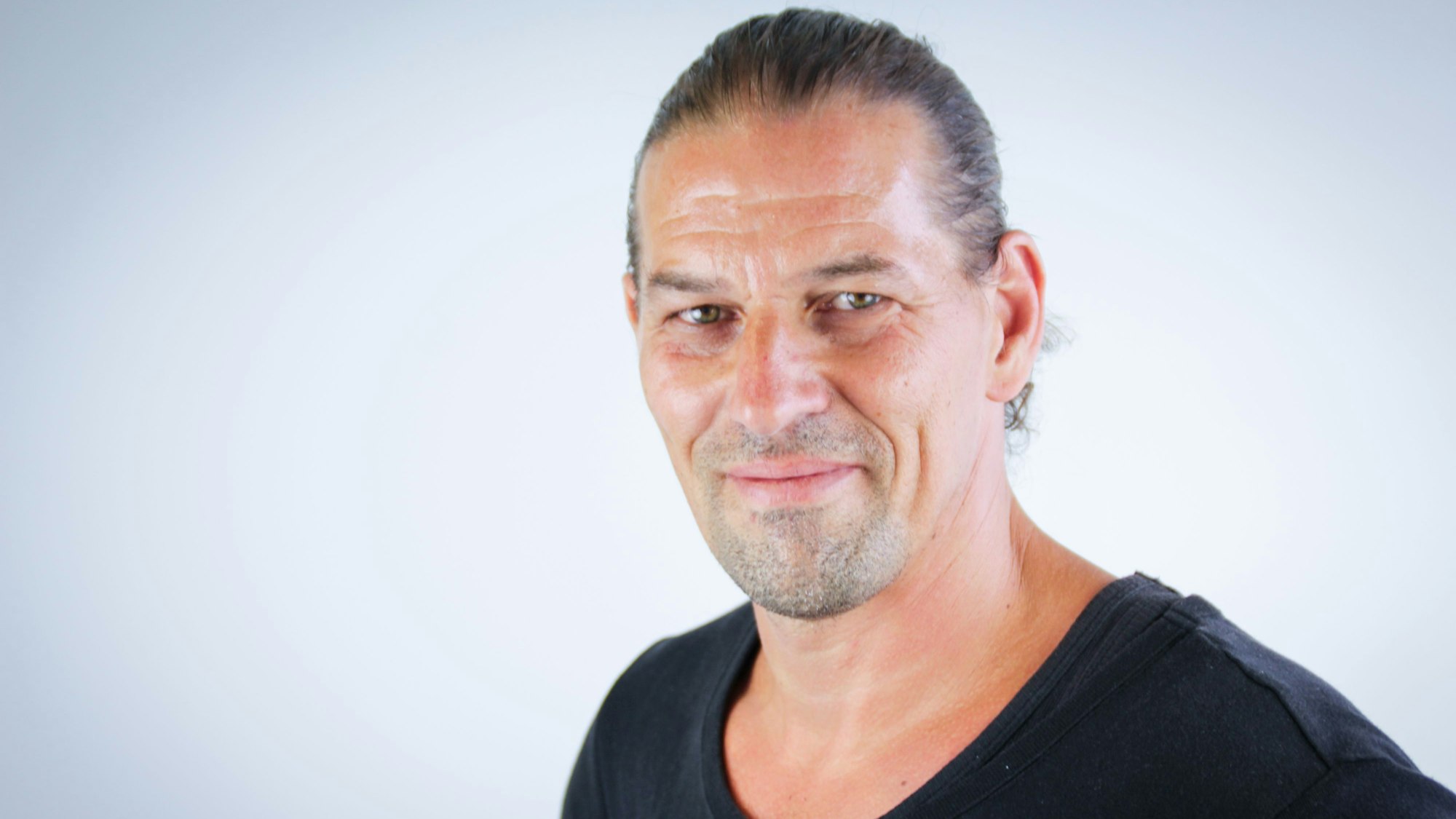 Ein Mann mit dunklem Pullover und dunklem zu einem Pferdeschwanz gebundenen Haar schaut lächelnd in die Kamera.