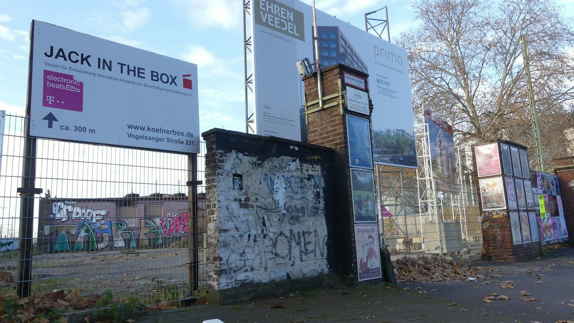 An einem Zaun hängen Schilder, die auf Jack in the box und das Neubauquartier Ehrenveedel hinweisen.