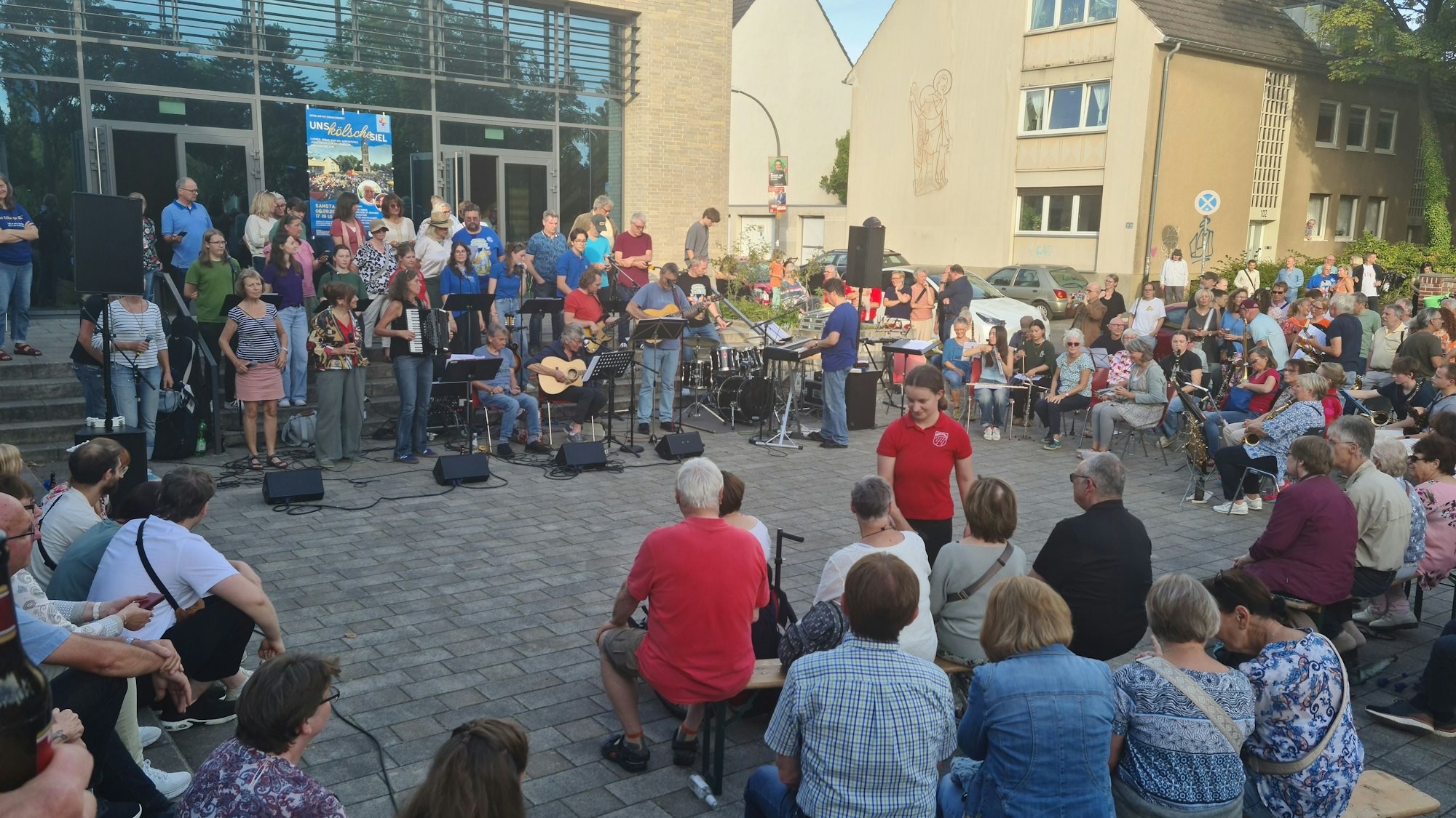 Auf einem Platz sind Musiker und viele Zuhörer versammelt, die auf Bänken sitzen.