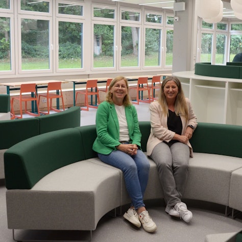 Zwei Frauen sitzen auf einem fast kreisrunden Sofa. Im Hintergrund stehen klassische Schulmöbel.