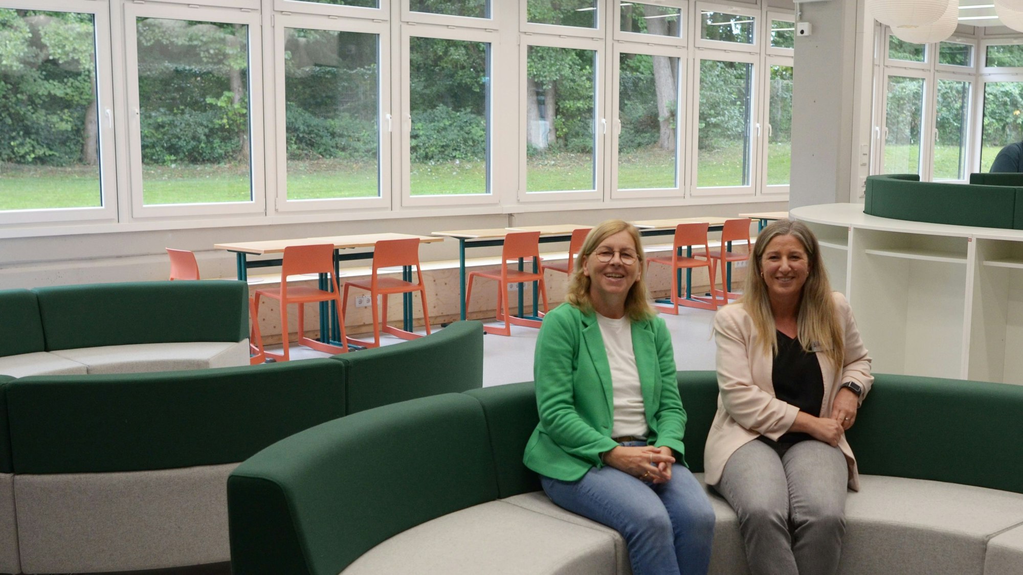 Zwei Frauen sitzen auf einem fast kreisrunden Sofa. Im Hintergrund stehen klassische Schulmöbel.