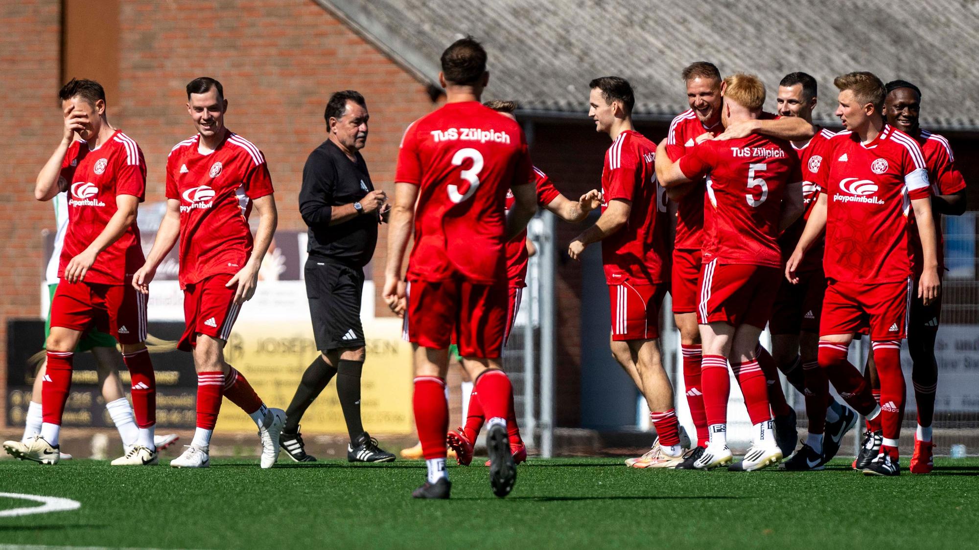 Zülpicher Spieler jubeln nach dem frühen 1:0. Ein Spieler am linken Bildrand hält sich ungläubig die Hand vors Gesicht. Der Schiedsrichter spricht mit den Spielern.