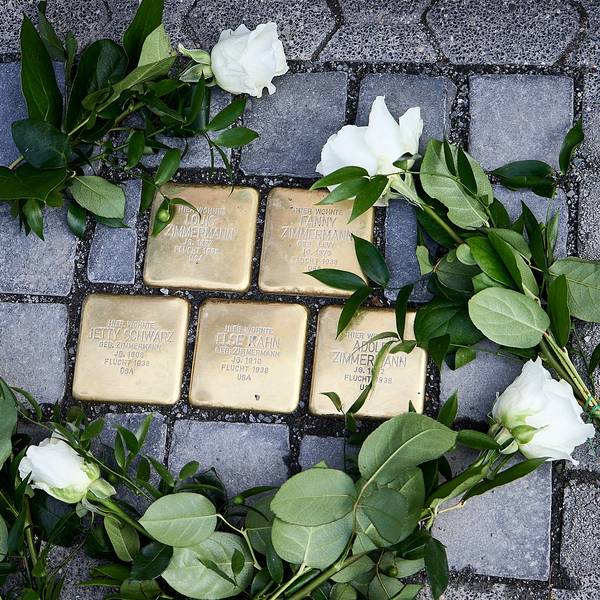 Das Bild zeigt fünf Pflastersteine aus Messing, auf denen Namen stehen. Drumherum liegen weiße Rosen.
