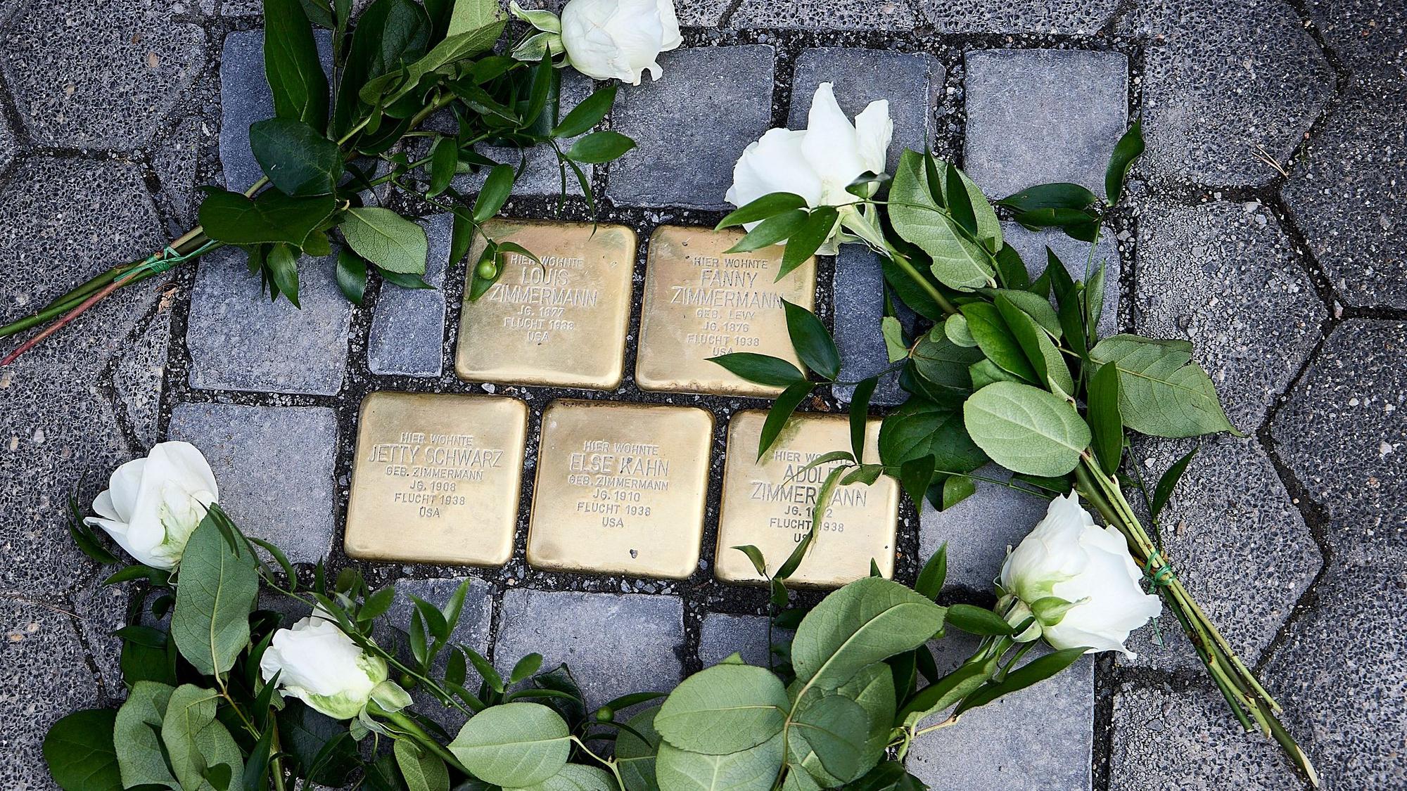 Das Bild zeigt fünf Pflastersteine aus Messing, auf denen Namen stehen. Drumherum liegen weiße Rosen.