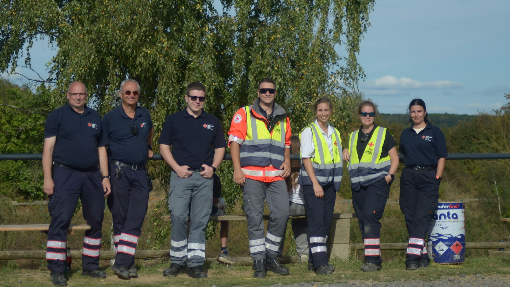 Vier Männer und drei Frauen in Rettungsdienstuniform stehen in einer Reihe an einem Zaun.