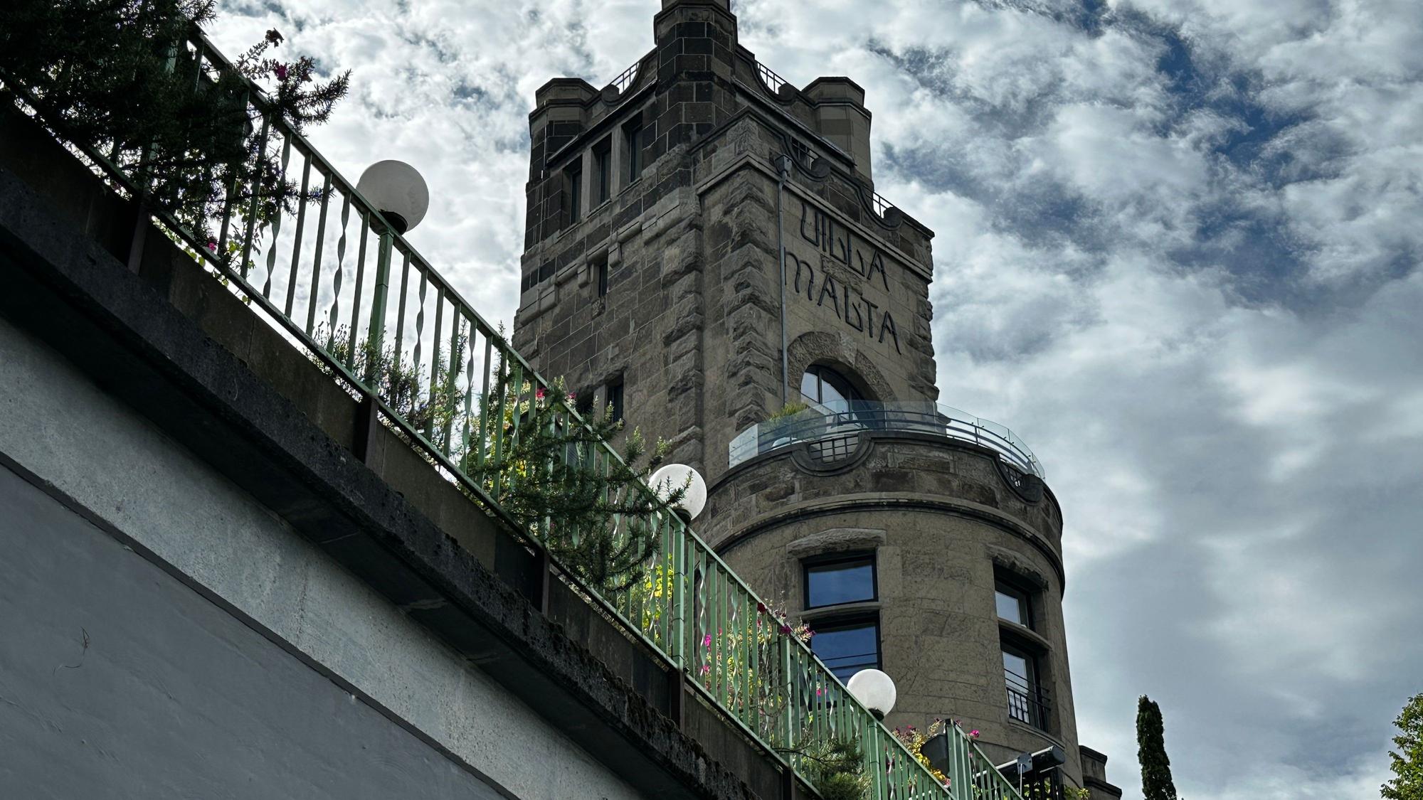Von unten fotografiert ist ein burgartiges Gebäude, das hinter einem Brückengeländer aufragt.