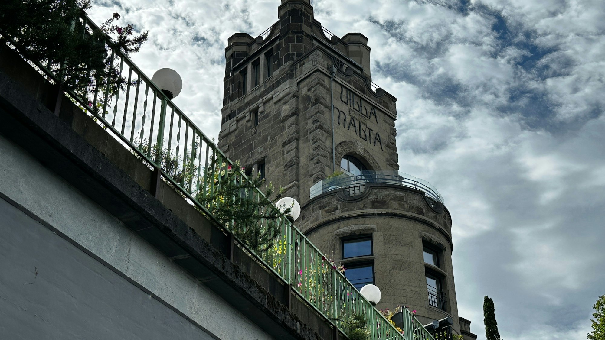 Von unten fotografiert ist ein burgartiges Gebäude, das hinter einem Brückengeländer aufragt.