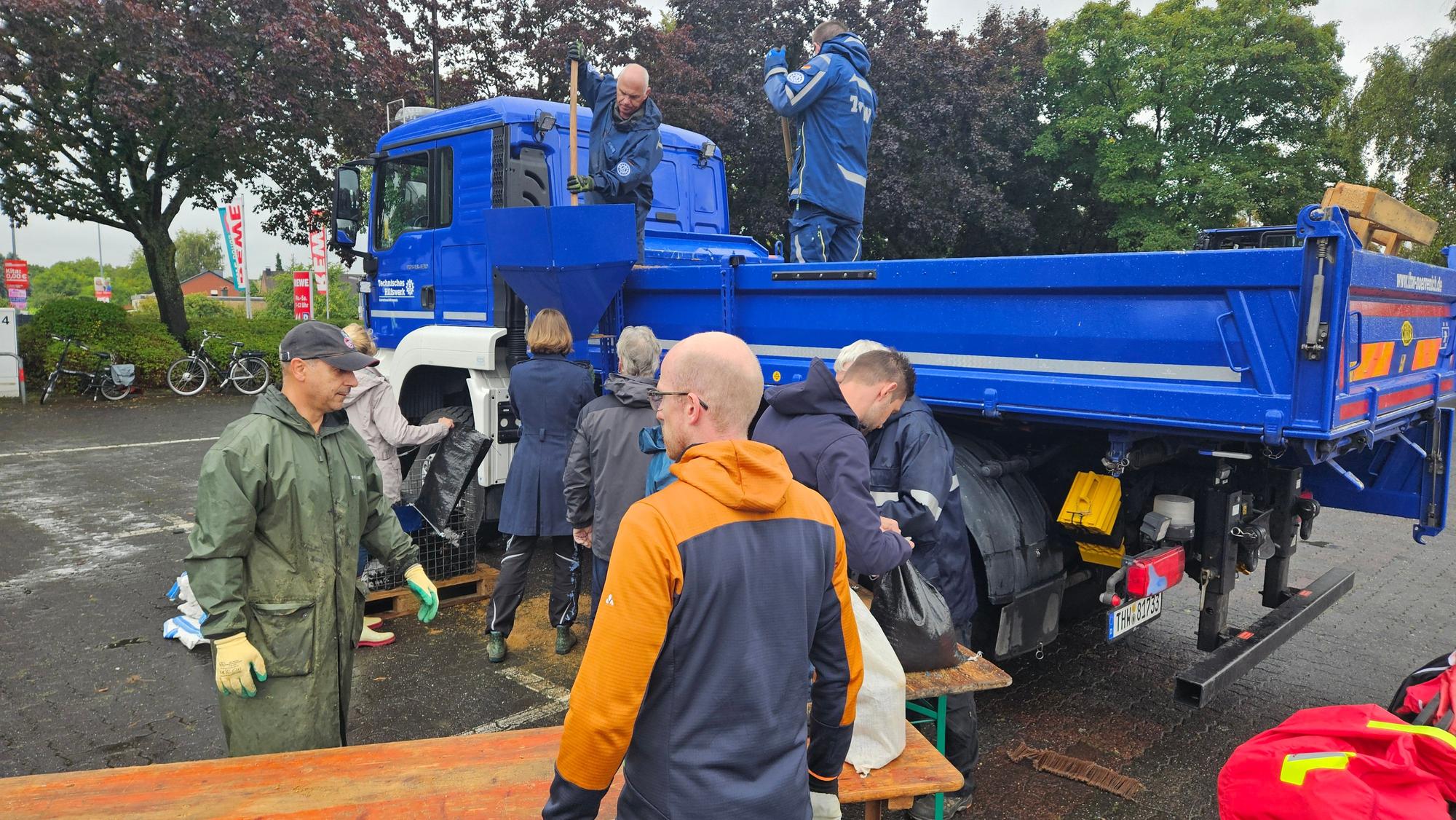 Auf dem Bild sind Helfer zu sehen, die Sandsäcke befüllen. Der Sand wird von einem blauen LKW herunter geladen.