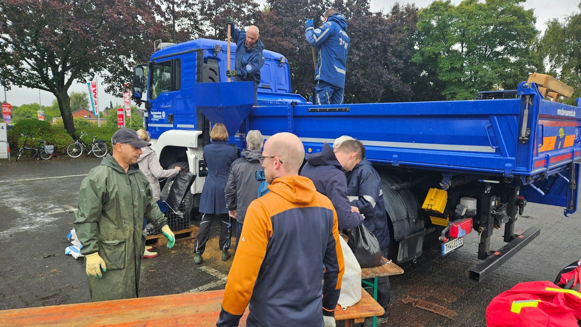 Auf dem Bild sind Helfer zu sehen, die Sandsäcke befüllen. Der Sand wird von einem blauen LKW herunter geladen.