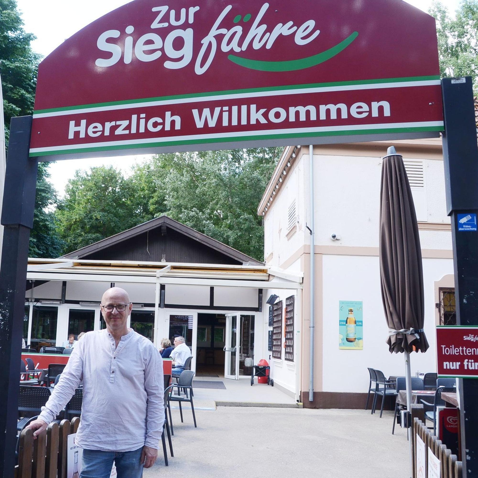 Ein Mann im weißen Hemd, mit Brille und Glatze steht vor einem Biergarten.