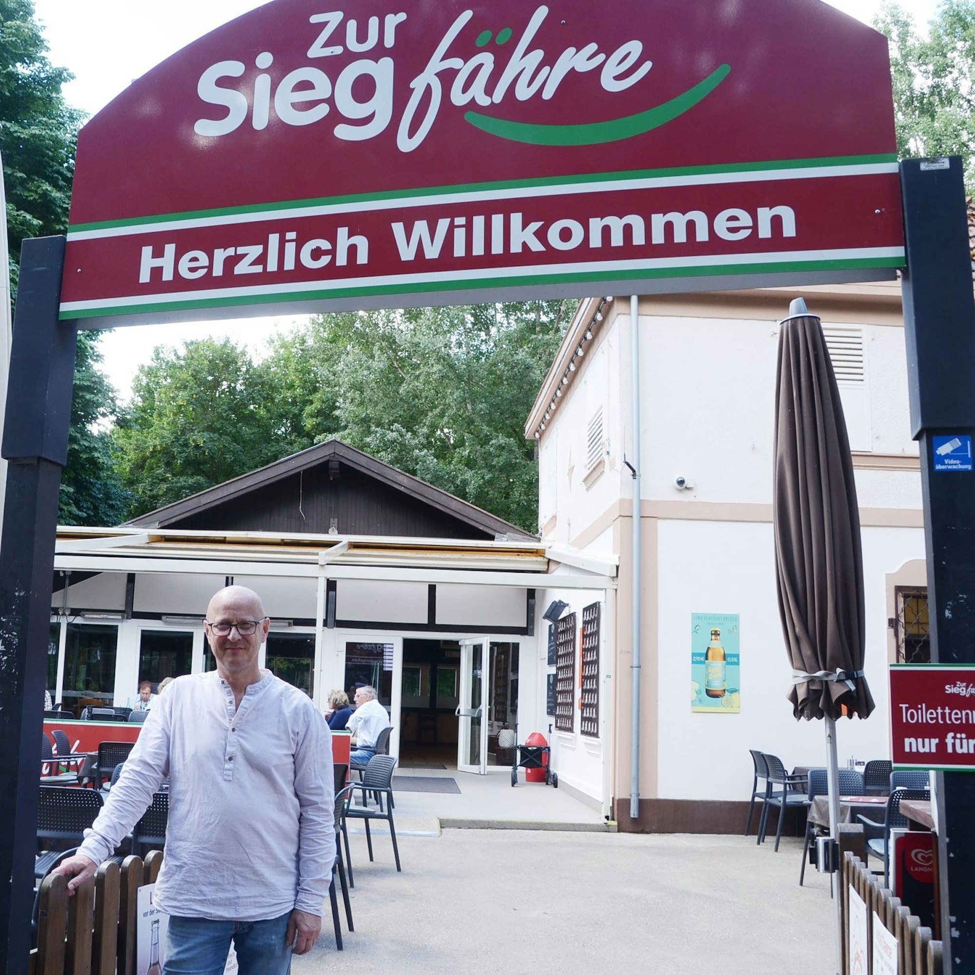 Ein Mann im weißen Hemd, mit Brille und Glatze steht vor einem Biergarten.