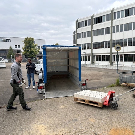 Das Foto zeigt, wie vor dem AOK-Gebäude Steine auf einer Palette mit dem Hubwagen auf einen Anhänger verladen werden.