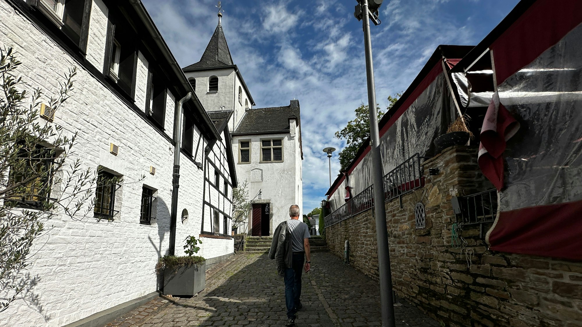 Ein Mann läuft eine Gasse hinab, links steht ein weißes Fachwerkhaus, das an eine Kapelle anschließt.