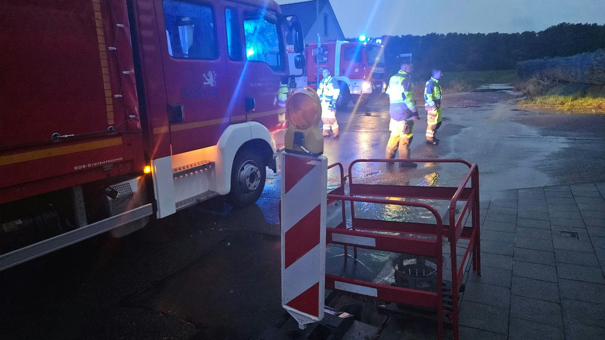 Zu sehen sind Feuerwehrleute und Feuerwehrfahrzeuge auf einer mit Wasser überfluteten Straße.
