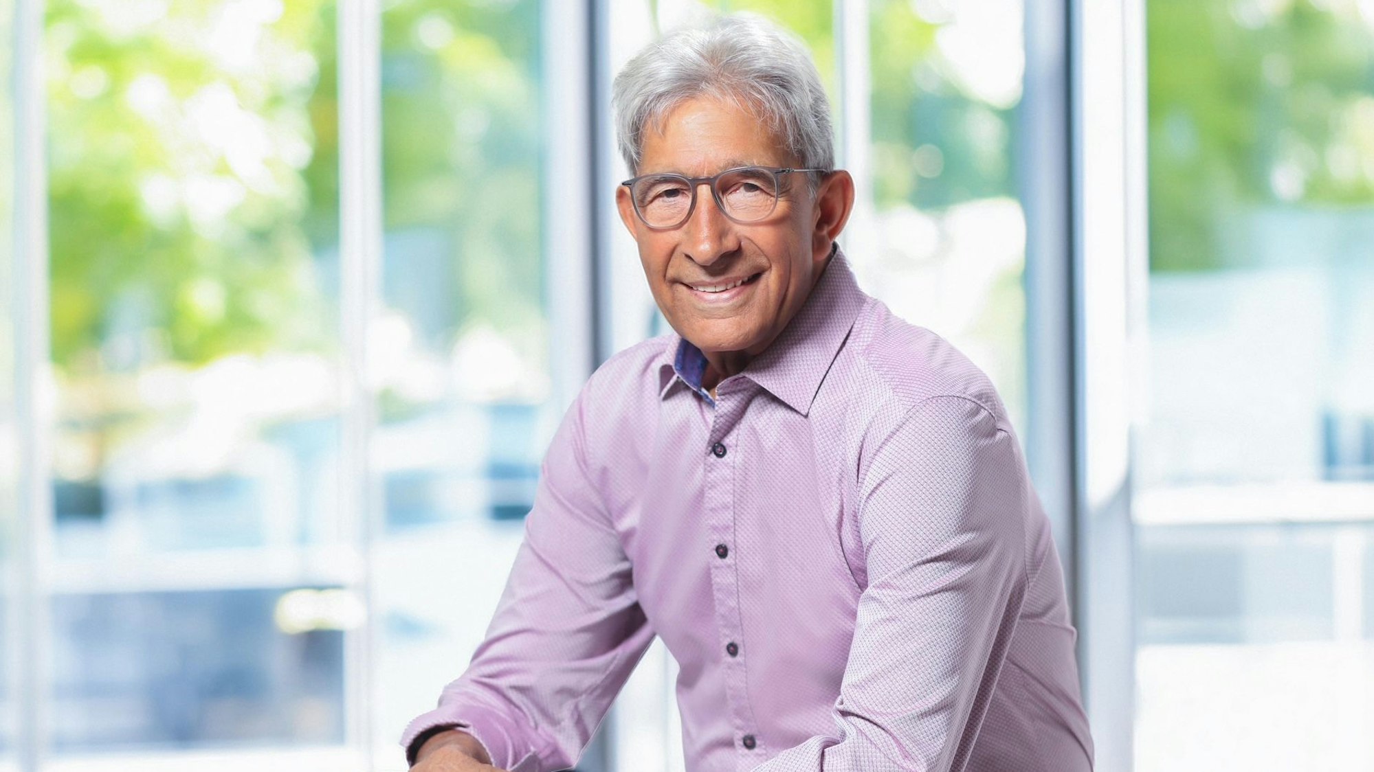 Der Großmeister Bajrush Saliu sitzt in einem lila Hemd vor einer Glaswand.