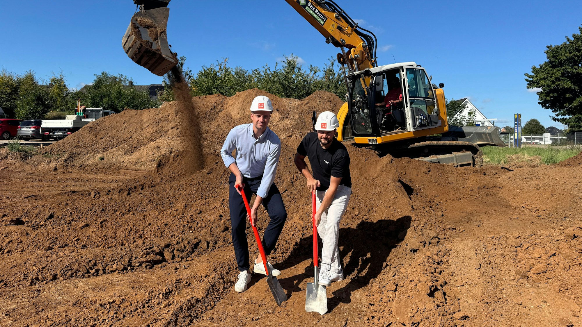 Das Bild zeigt die beiden Geschäftsführer beim symbolischen Spatenstich. Im Hintergrund steht ein Bagger.