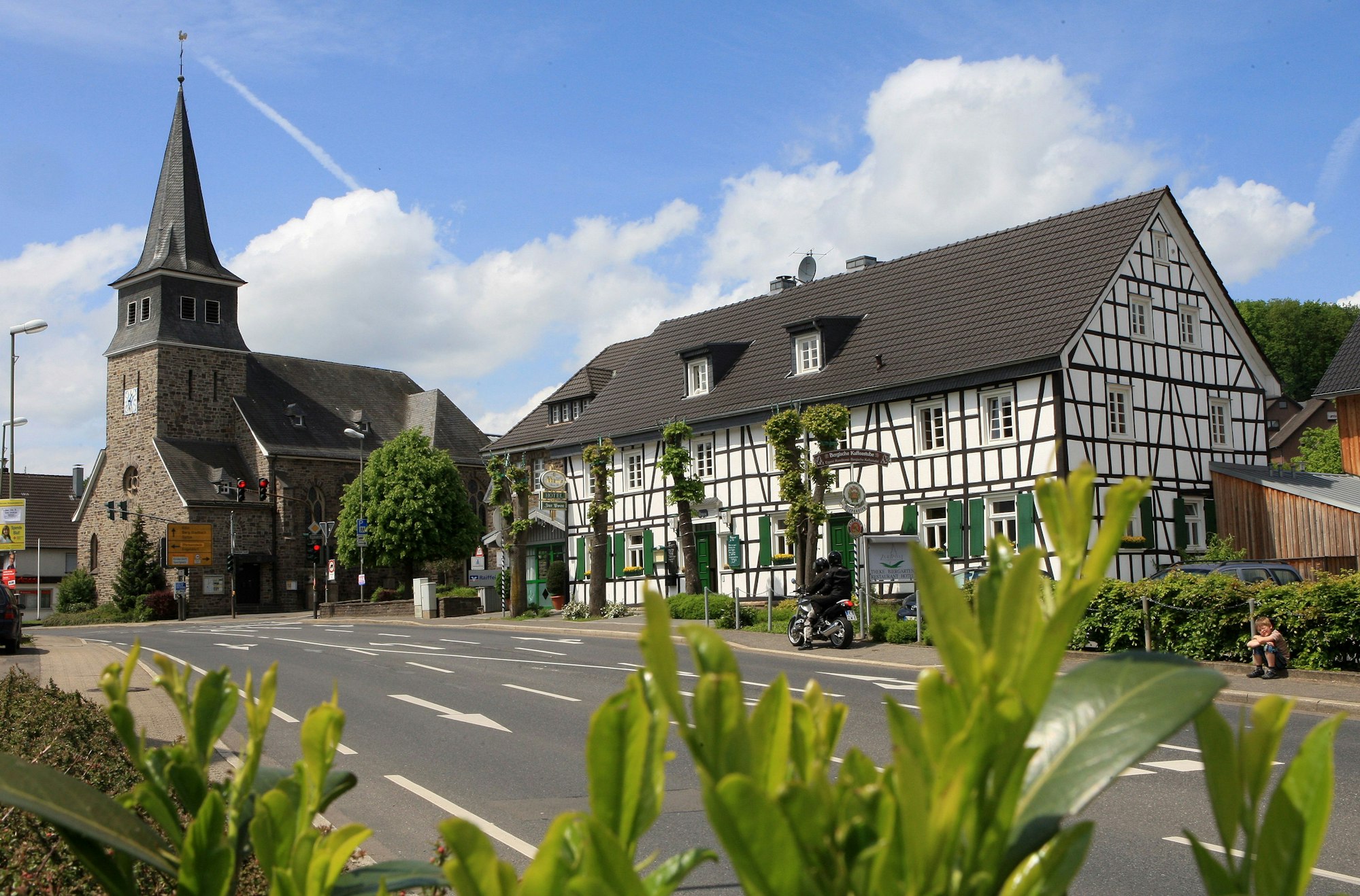 Eine Kirche mit trutzigem Turm aus Bruchstein. Daneben ein historisches Fachwerkhaus. Davor eine Straße.