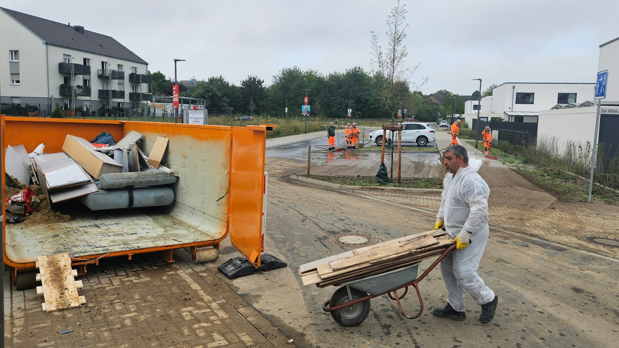 Zu sehen ist, wie ein Mann in einem weißen Overall Sperrmüll auf einer Schubkarre in einen Container schiebt.