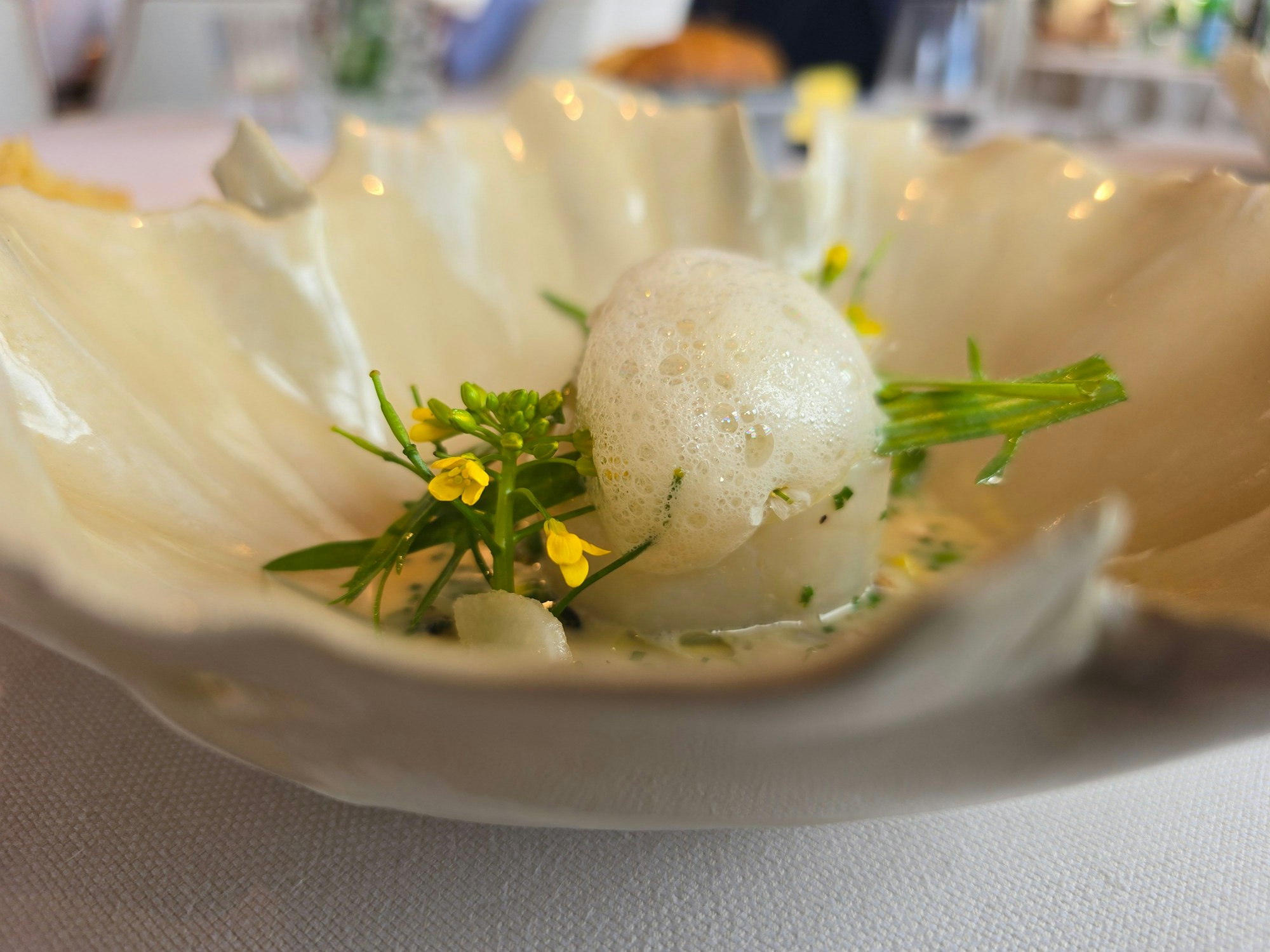 Ein Teller in Blattform mit einem feinen Arrangement aus Fisch, Schaum und frischer Blüte ist zu sehen.