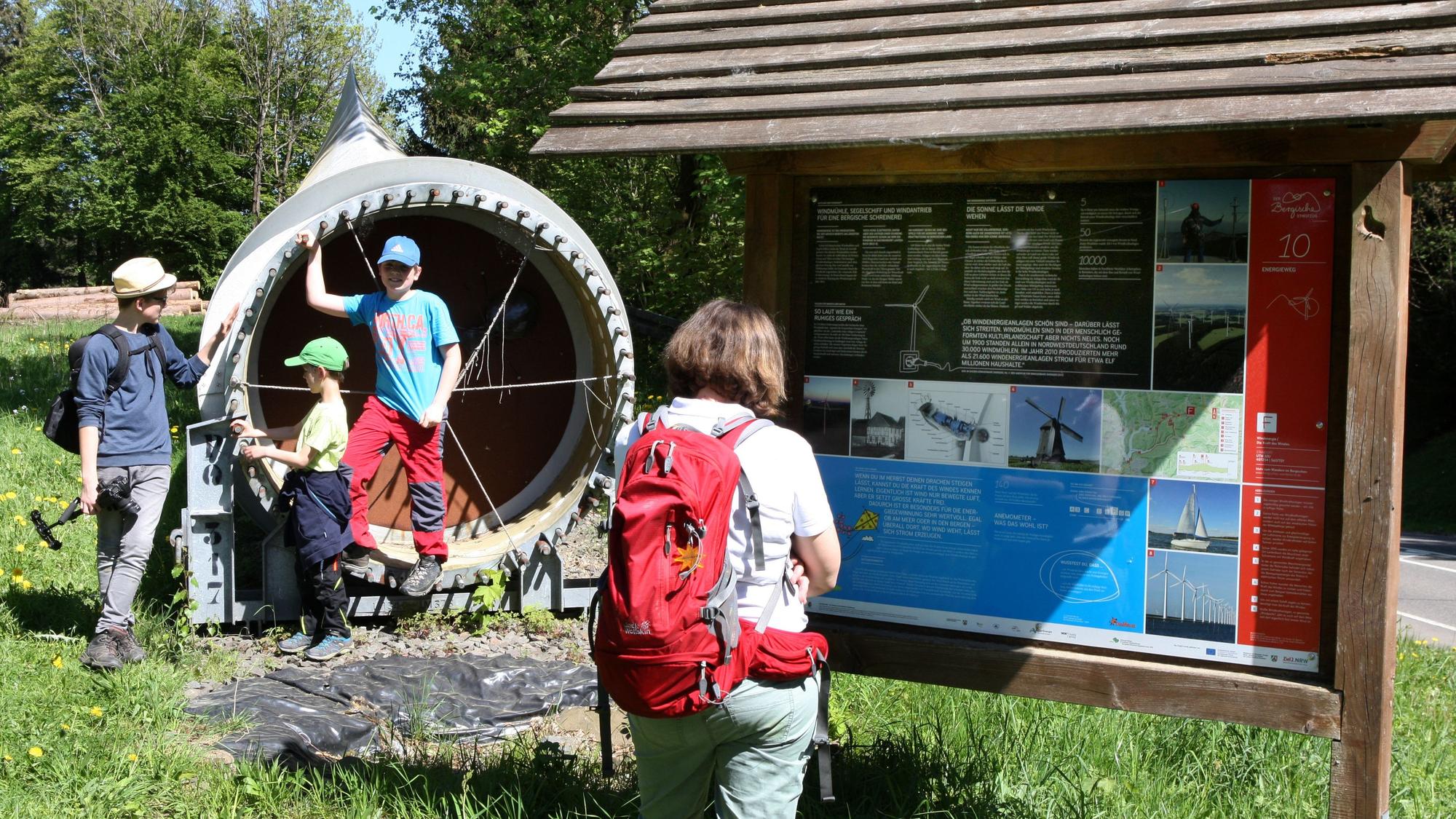 Ein Rotorflügel einer Windkraftanlage liegt neben einer Infotafel des Energieweges bei Gummersbach. Die Texte der Tafel liest eine Frau, am Rotorflügel spielen Kinder.