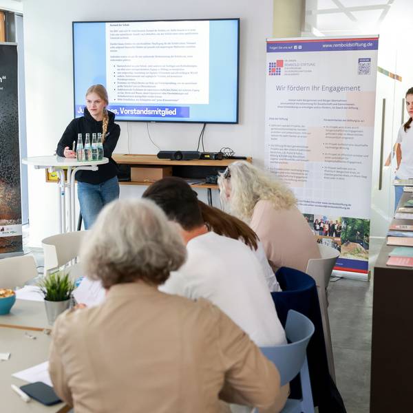 Mehrere junge Leute stehen vor einer Leinwand, eine junge Frau steht an einem Stehtisch und spricht.
