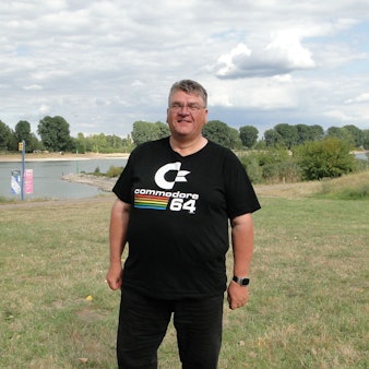 Ein Mann mit Brille und schwarzer Hose und Shirt steht auf einer Wiese am Rhein.