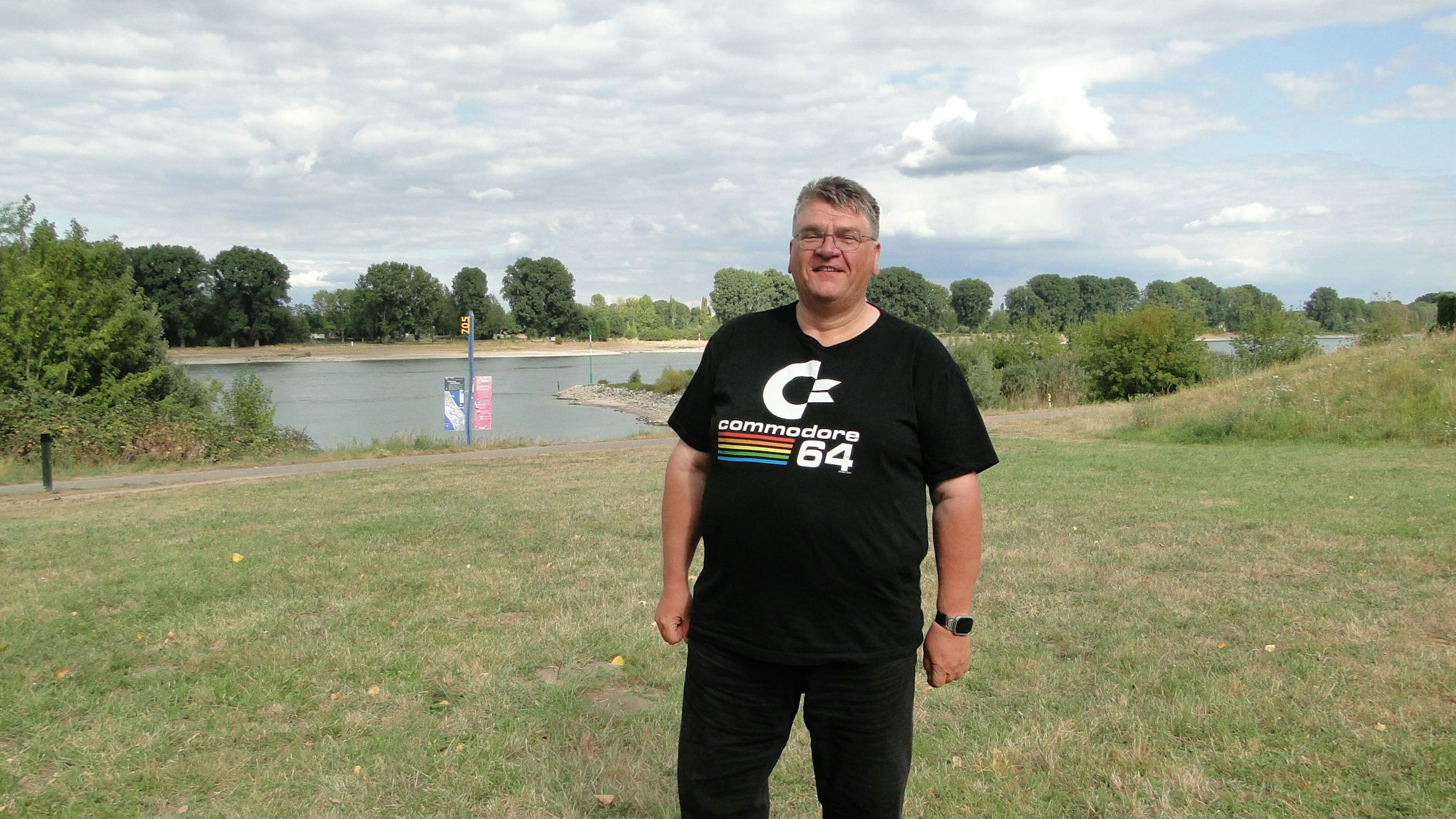 Ein Mann mit Brille und schwarzer Hose und Shirt steht auf einer Wiese am Rhein.