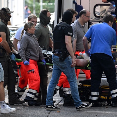 Einsatzkräfte bringen die Trage mit einer verletzten, tatverdächtigen Person zu einem Rettungswagen. Kurz nach dem Messerangriff auf eine Lehrerin an einem Berufskolleg in Essen ist die tatverdächtige Person gefasst worden.
