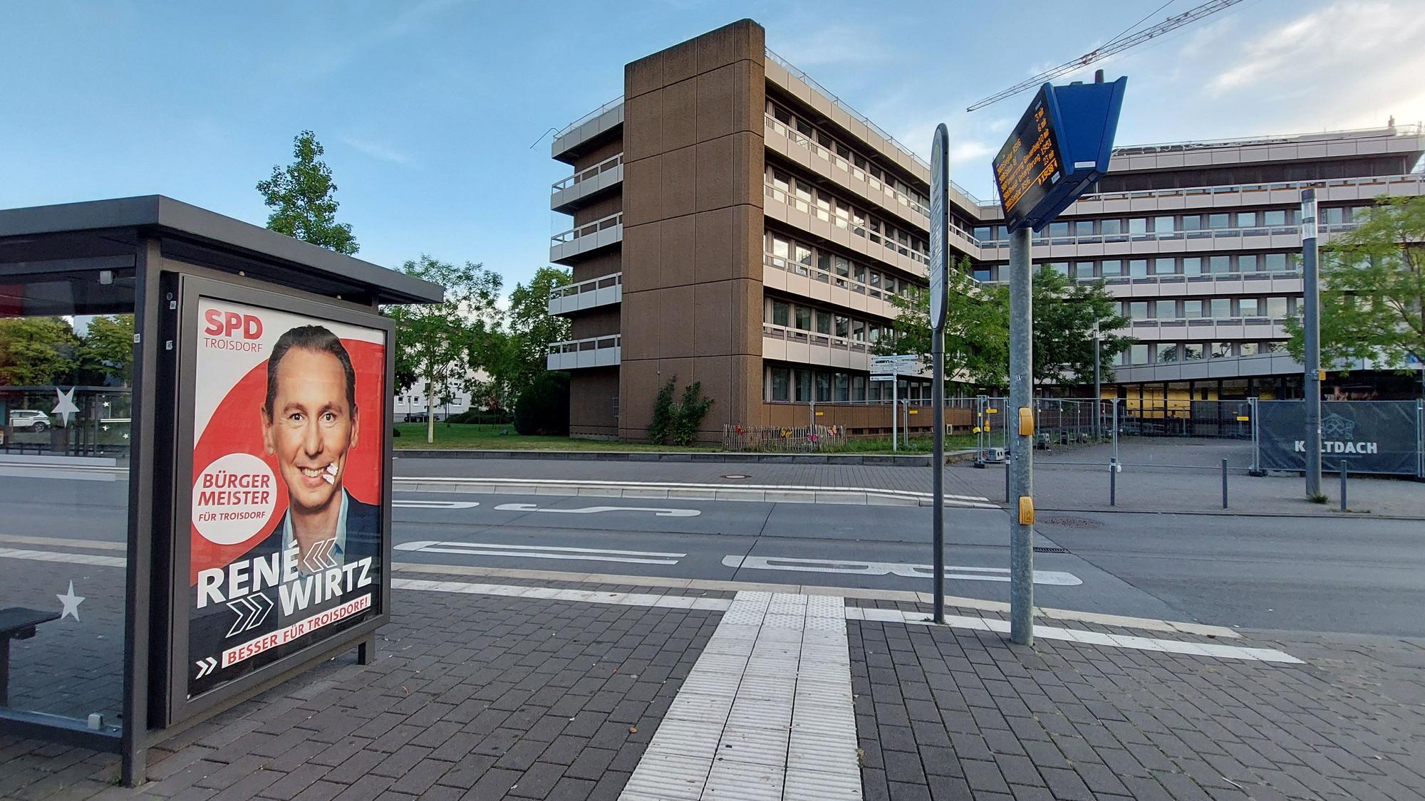 Werbeplakat für einen SPD-Kandidaten an einer Bushaltestelle gegenüber einem Verwaltungsgebäude.