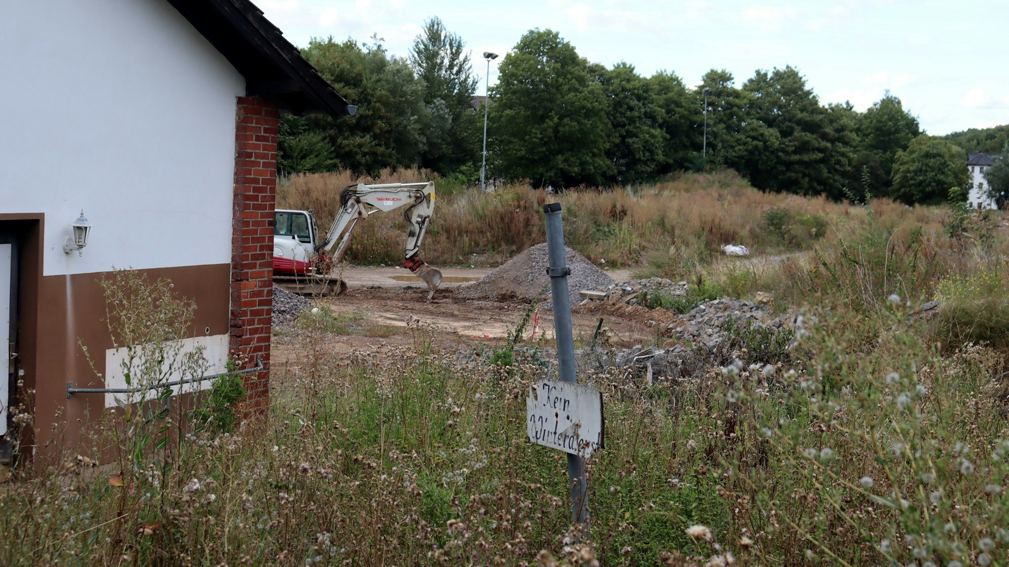Der Sportplatz in Bad Münstereifel besteht aus Geröllhaufen und ist zugewuchert. Im Hintergrund steht ein Bagger, im Vordergrund ist das schon vor der Flut nicht mehr nutzbare Vereinsheim zu sehen, daneben steht ein Schild mit dem Hinweis „Kein Winterdienst“.