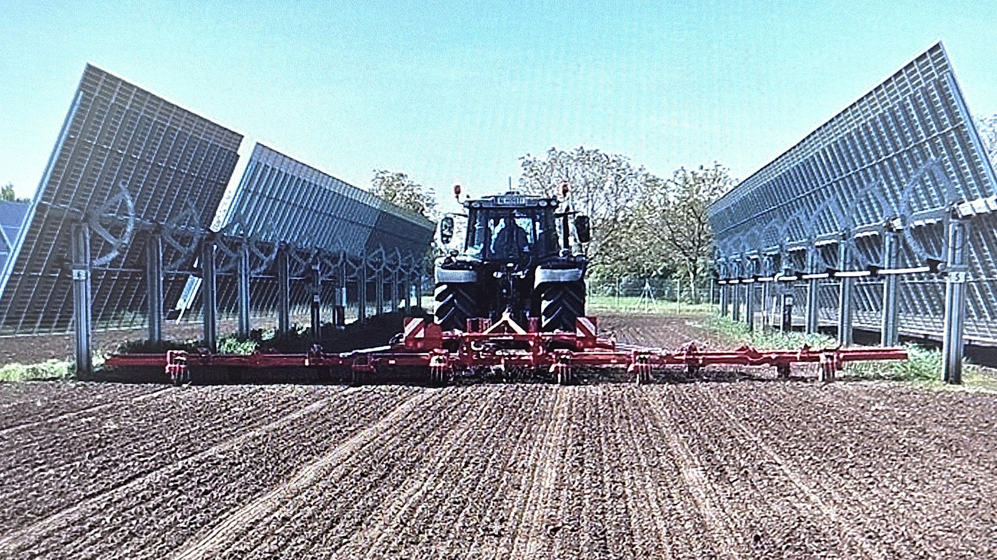 Ein Traktor mit angehängter Maschine fährt zwischen zwei Modulreihen hindurch.