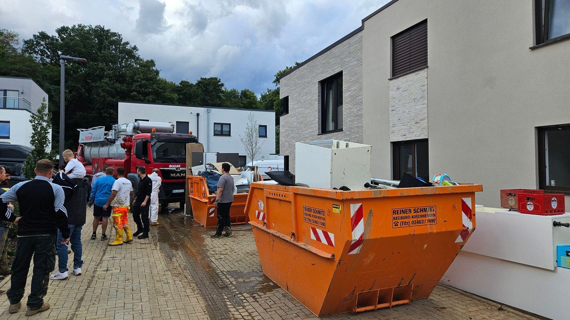 Zu sehen sind Container voller Sperrmüll in einer Naubausiedlung, Menschen stehen auf der Straße.