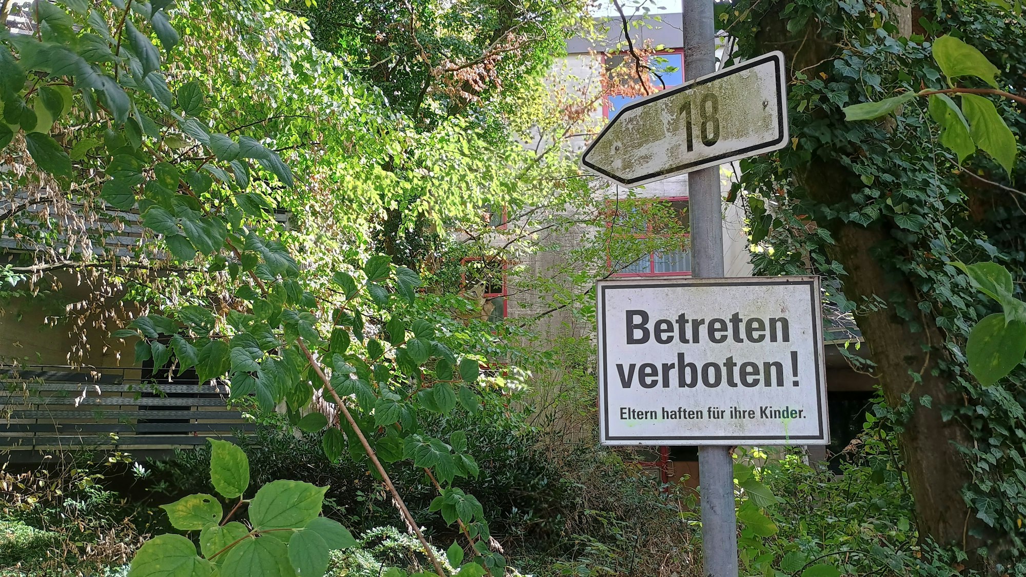 Ein Schild mit der Nummer 18, darunter ein Schild mit der Aufschrift: „Betreten verboten! Eltern haften für ihre Kinder“. Im Hintergrund das Gebäude, stark von Pflanzen verdeckt.