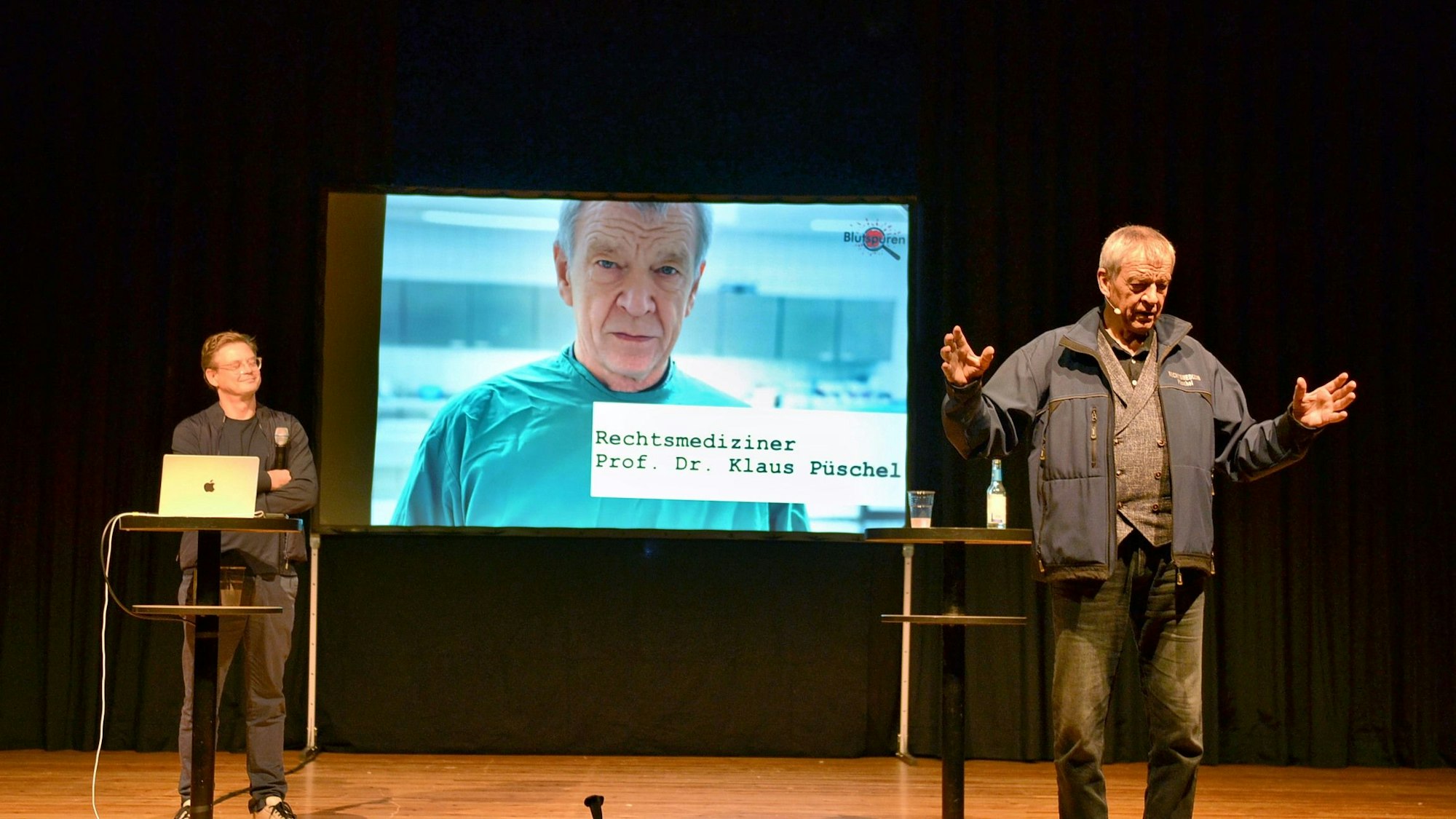Prof. Dr. Klaus Püschel steht rechts auf der Bühne und gestikuliert. Auf einer Leinwand ist ein großes Porträt von ihm zu sehen. Links steht Moderator Dr. Tino Grosche.
