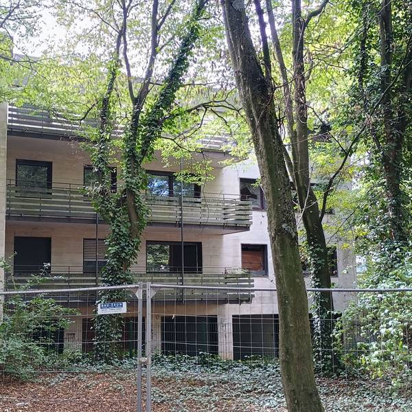 Das Bild zeigt das umzäunte Gebäude des Wohnparks. Viele Fenster sind eingeschlagen, einige Bäume verdecken die Sicht auf das Haus.