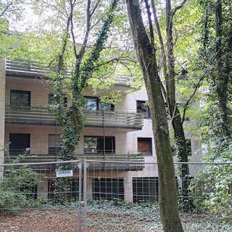 Das Bild zeigt das umzäunte Gebäude des Wohnparks. Viele Fenster sind eingeschlagen, einige Bäume verdecken die Sicht auf das Haus.