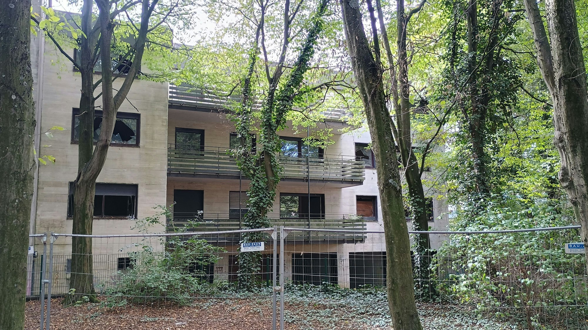 Das Bild zeigt das umzäunte Gebäude des Wohnparks. Viele Fenster sind eingeschlagen, einige Bäume verdecken die Sicht auf das Haus.