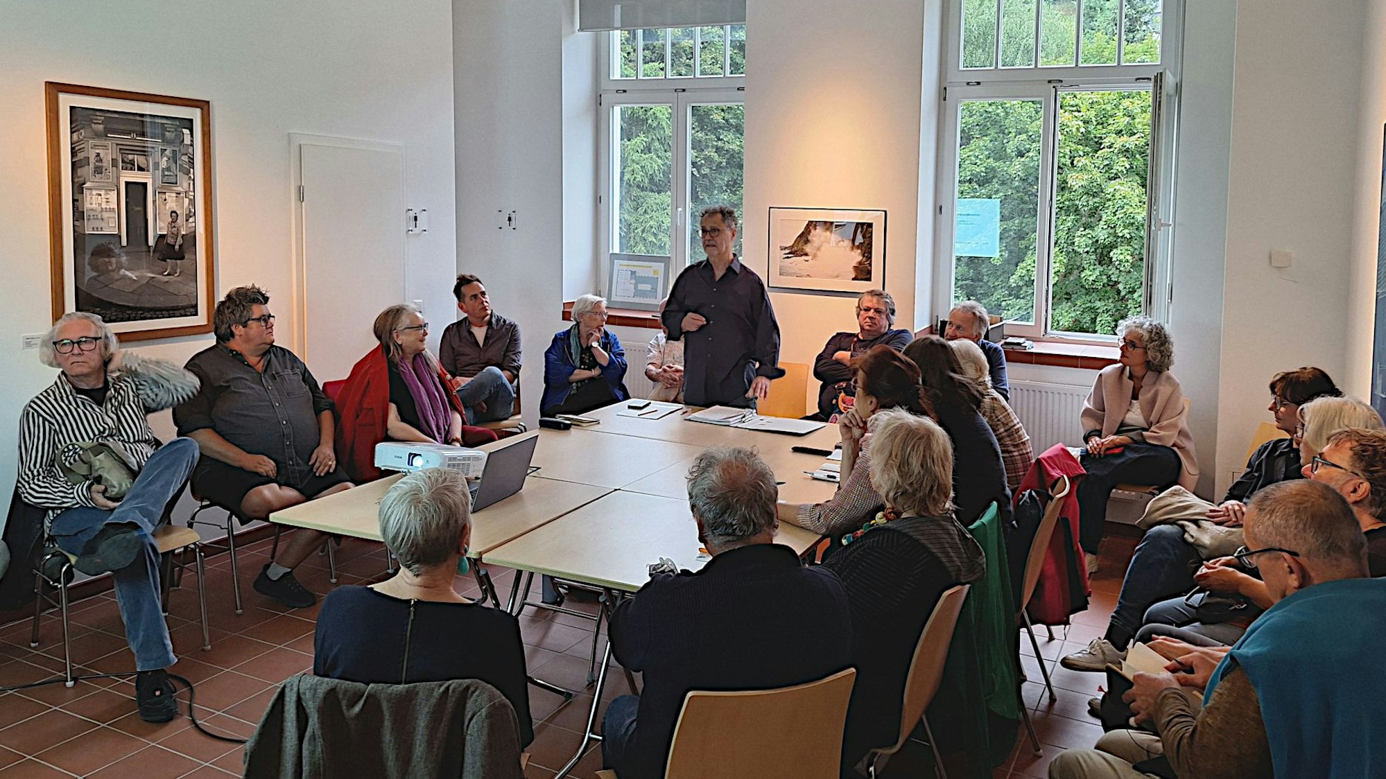 Menschen sitzen um einen Tisch herum. Ein Mann steht am Tisch und redet.