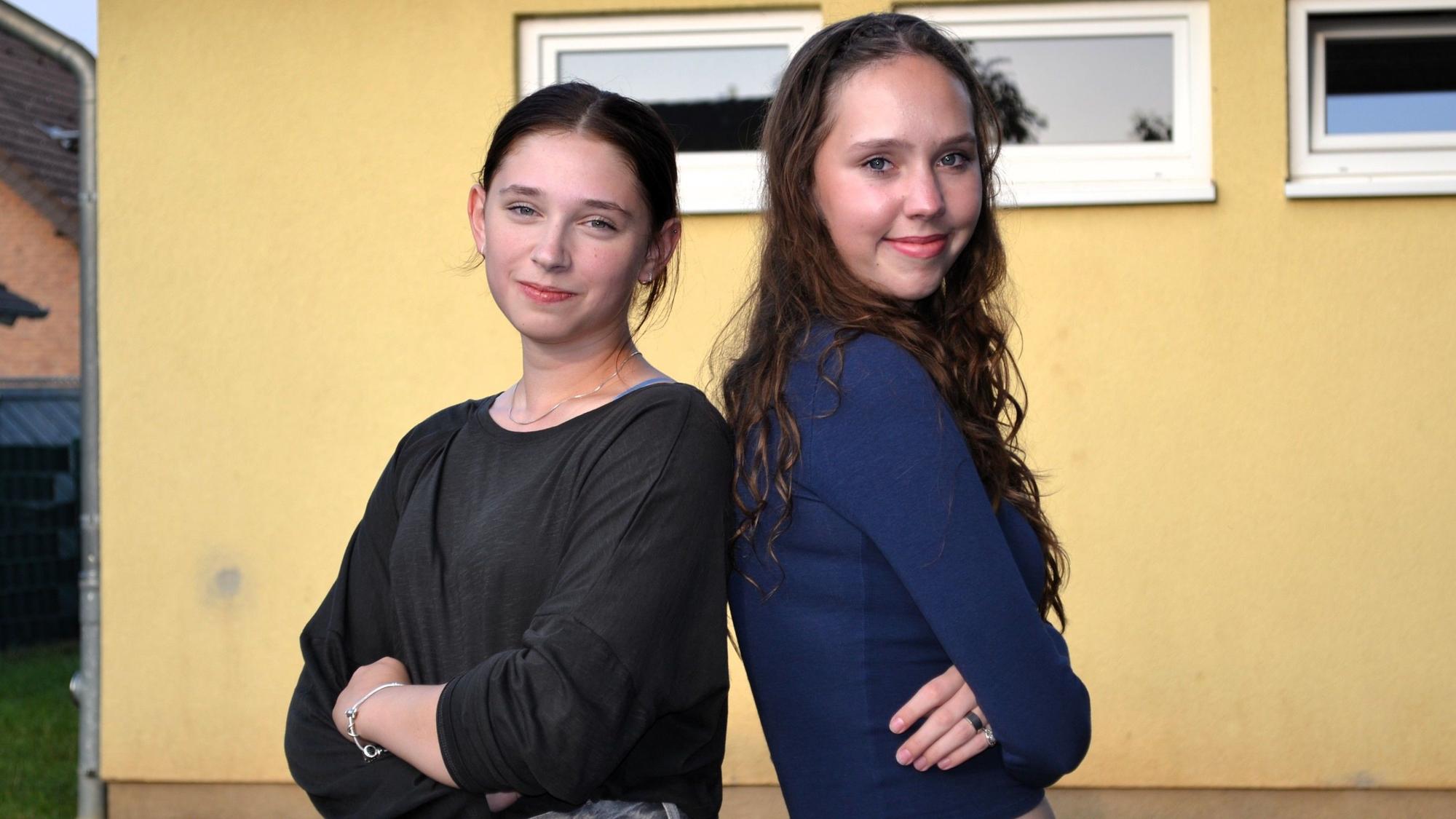 Zwei junge Mädchen stehen Rücken an Rücken und schauen in die Kamera.