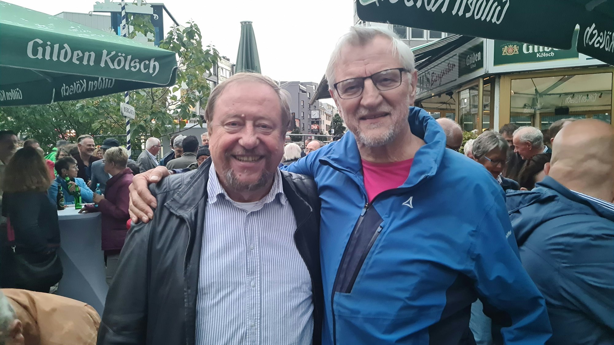 Ein Mann im hellblau-weiß gestreiftem Hemd und schwarzer Jacke steht neben einem Mann mit Brille im blauen Anorak.