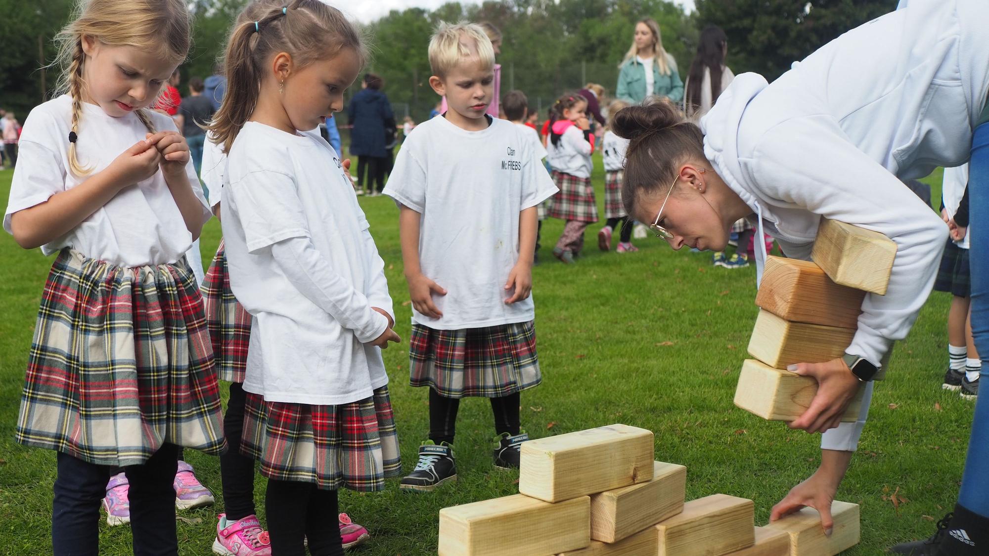 Zu sehen sind drei Kinder, die Schottenrock tragen und Holzklötze stapeln.
