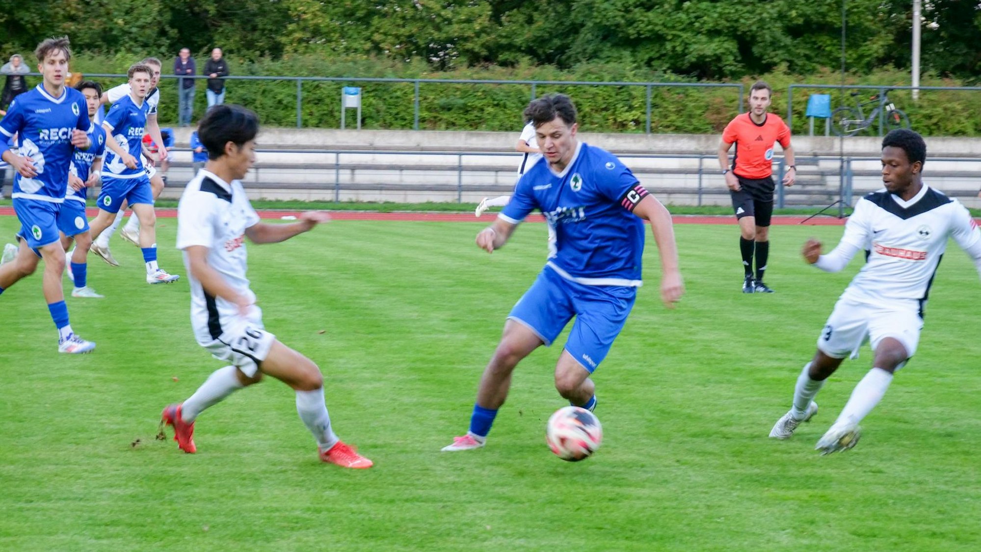 Das Foto zeigt einen blau gekleideten Königsdorfer Fußballer, der mit dem Ball am Fuß eine Anspielstation sucht, doch zwei weiße Frechener stellen den Raum zu.