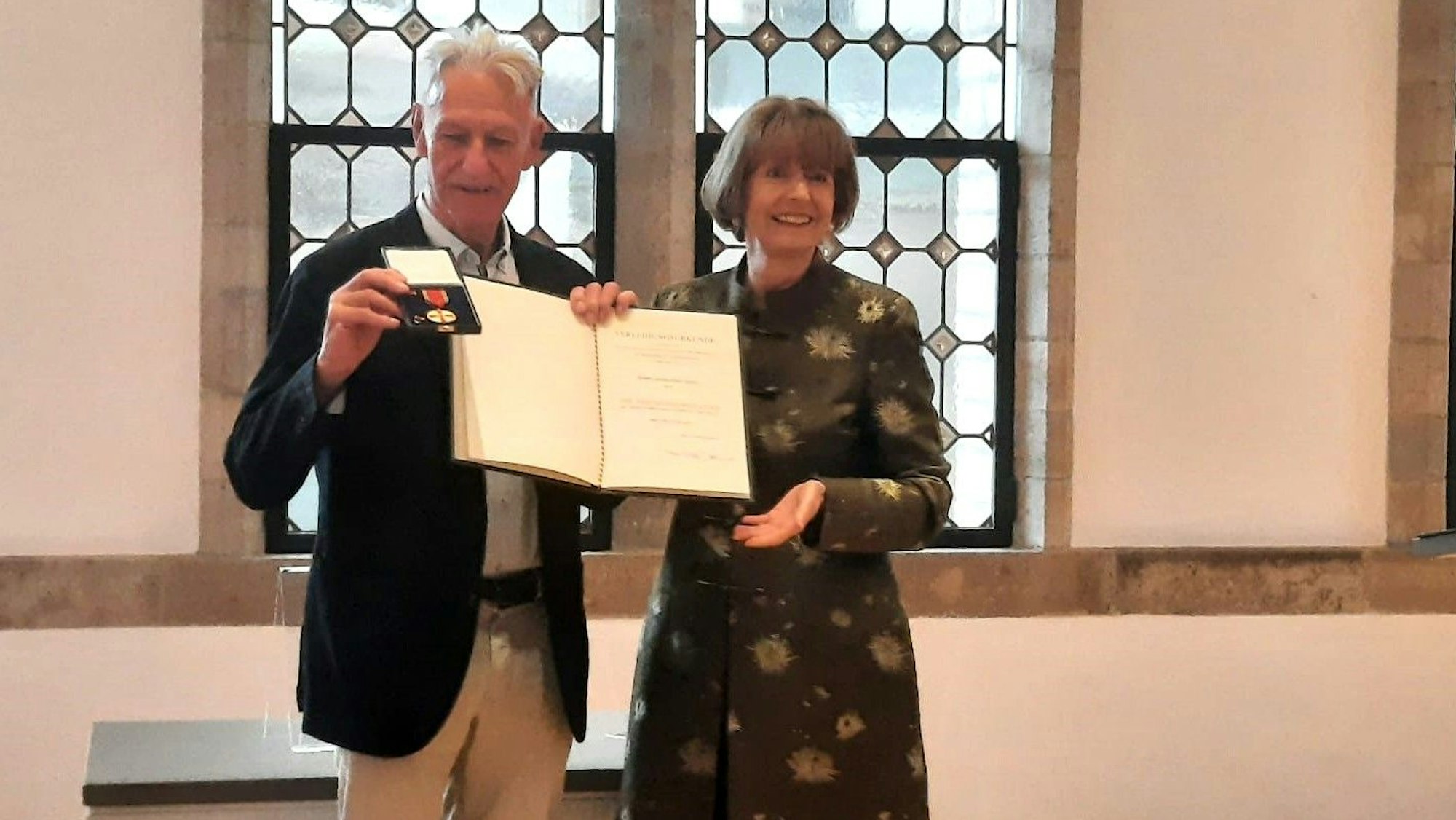 Ein äterer Mann mit grauem Haar hält ein Buch geöffnet vor sich, neben ihm steht eine elegante Frau im braunen Kleid, Henriette Reker.