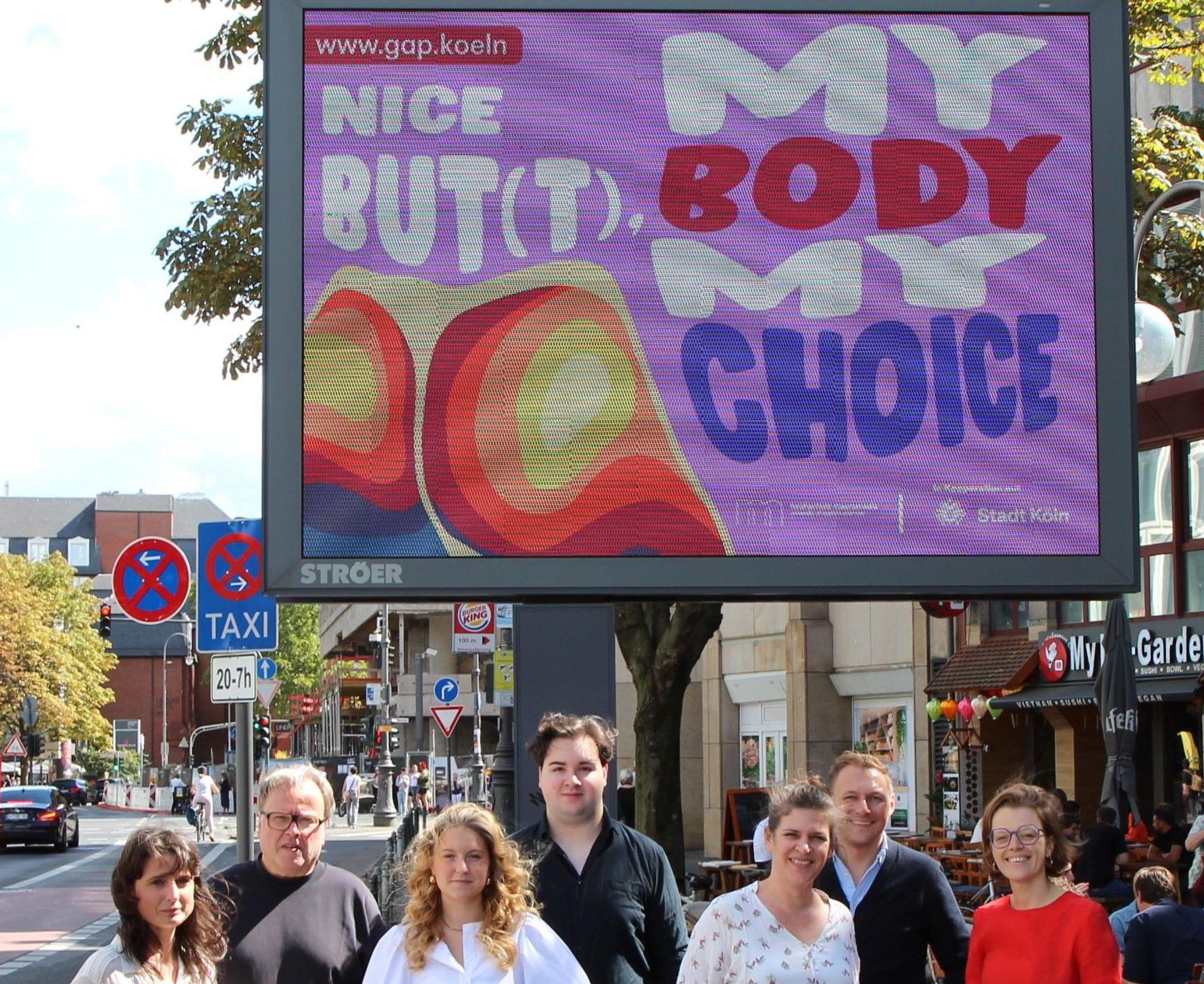 Die Beteiligten der "My Body My Choice" Kampagne stehen unter einer digitalen Werbetafel, wo die Kampagne abgedruckt ist