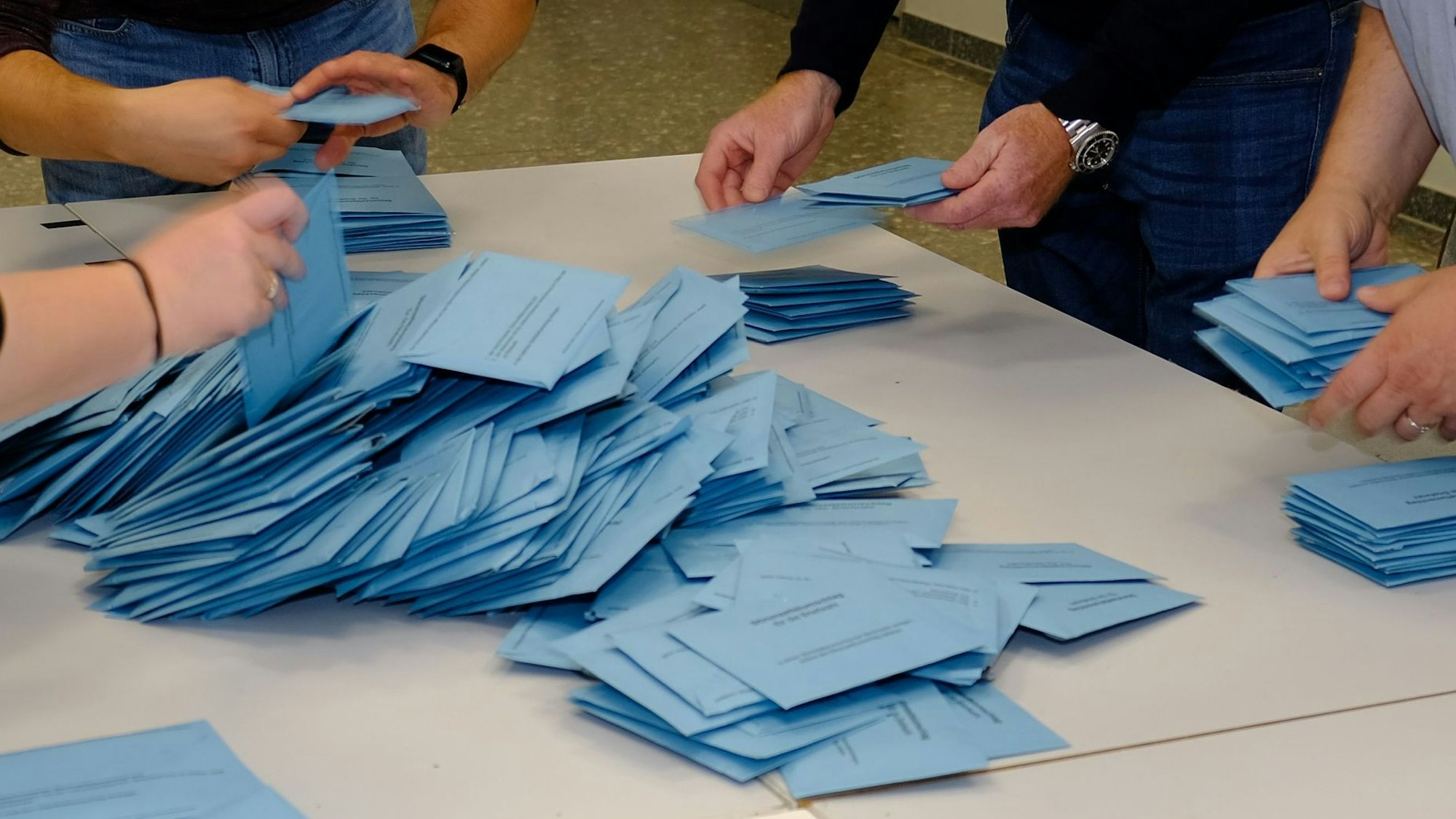 Vier Menschen greifen mit den Händen nach blauen Stimmzetteln bei der Kommunalwahl in Mechernich, die zur Auszählung auf einem Tisch liegen.
