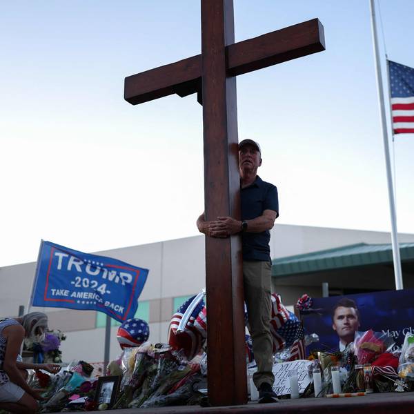 TOPSHOT - A US flag flies half-staff as a man carries a large cross near a makeshift memorial for Turning Point USA Founder Charlie Kirk outside of the Turning Point USA headquarters in Phoenix, Arizona, on September 12, 2025. The widow of prominent right-wing activist Charlie Kirk pledged on September 12 to carry on her husband's work, after US authorities announced his alleged assassin had finally been captured. The 31-year-old Kirk was hit by a single bullet while addressing a large crowd at Utah Valley University in the town of Orem on September 10. (Photo by CHARLY TRIBALLEAU / AFP)