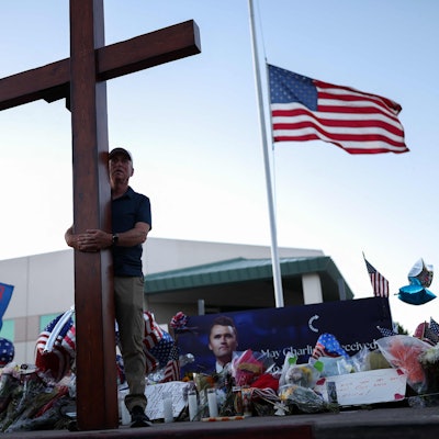 TOPSHOT - A US flag flies half-staff as a man carries a large cross near a makeshift memorial for Turning Point USA Founder Charlie Kirk outside of the Turning Point USA headquarters in Phoenix, Arizona, on September 12, 2025. The widow of prominent right-wing activist Charlie Kirk pledged on September 12 to carry on her husband's work, after US authorities announced his alleged assassin had finally been captured. The 31-year-old Kirk was hit by a single bullet while addressing a large crowd at Utah Valley University in the town of Orem on September 10. (Photo by CHARLY TRIBALLEAU / AFP)